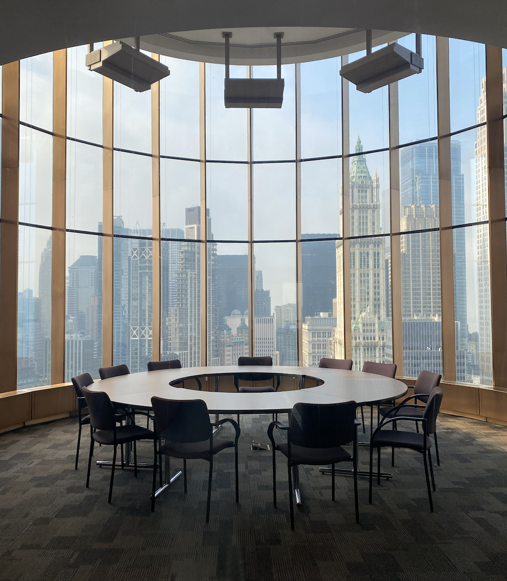 A roundtable and chairs in an office space with floor-to-ceiling windows overlooking the New York City skyline.